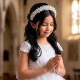 Una niña de cabello oscuro sonríe suavemente en una iglesia, usando una diadema ancha de comunión decorada con densos racimos de perlas blancas sobre un vestido de encaje - VII Sacramentos Colombia
