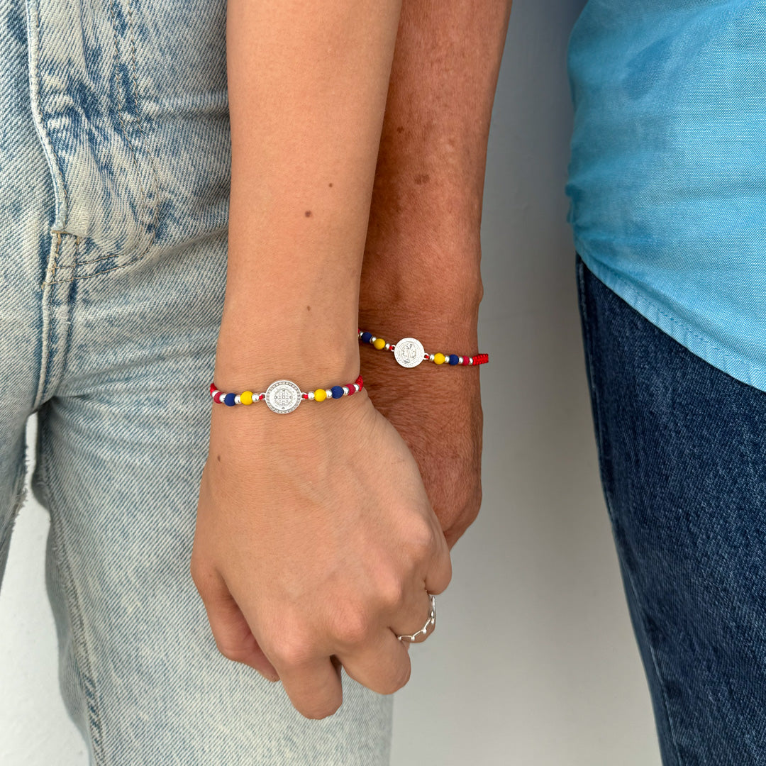 Dos personas tomadas de la mano usando la pulsera tricolor Colombia de hilo rojo con medalla de plata de edición especial.
