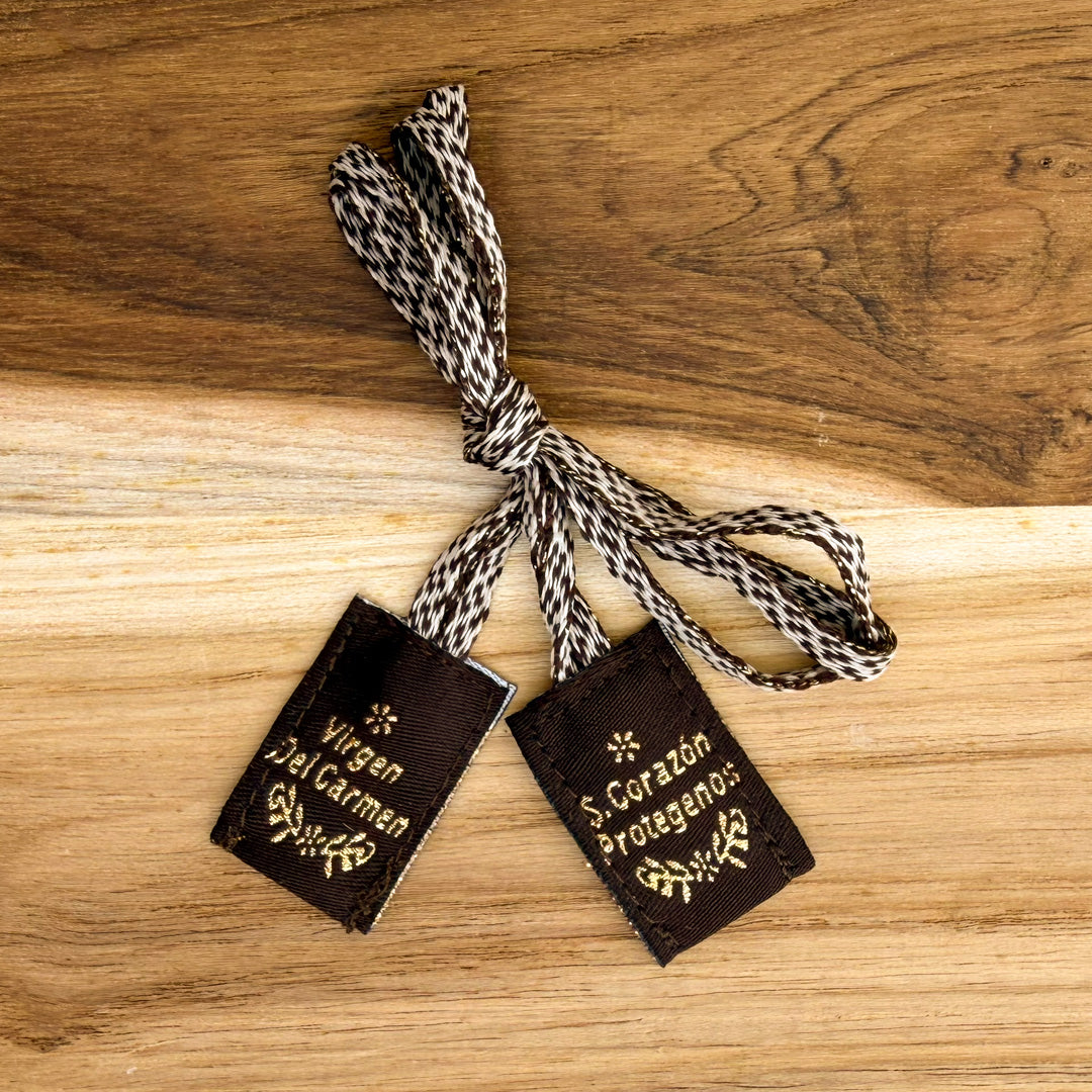 Reverso de escapulario bordado con letras en hilo dorado que dicen Virgen del Carmen y S. Corazón Protégenos sobre tela café.
