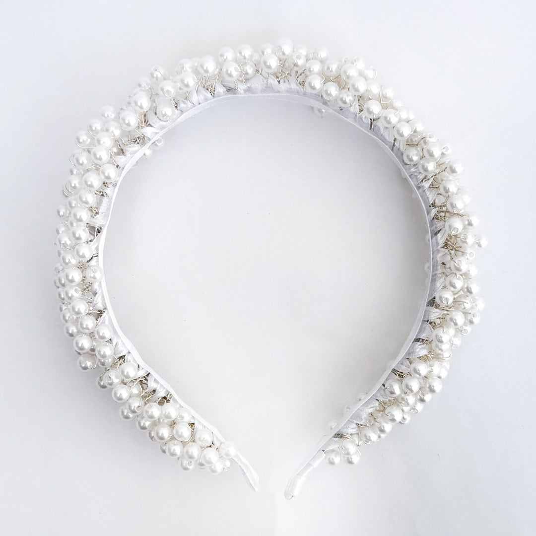 Una niña de cabello oscuro sonríe suavemente en una iglesia, usando una diadema ancha de comunión decorada con densos racimos de perlas blancas sobre un vestido de encaje - VII Sacramentos Colombia
